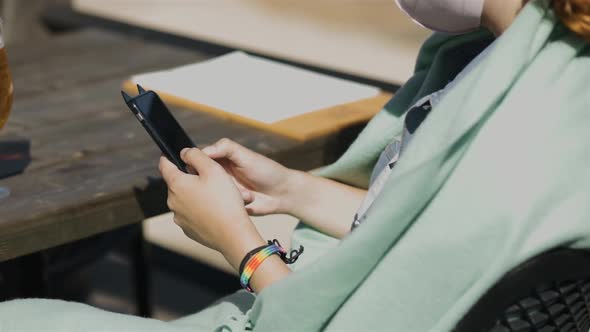 LGBT Woman Uses Smartphone in Cafe Wearing Rainbow Bracelet and Face Mask