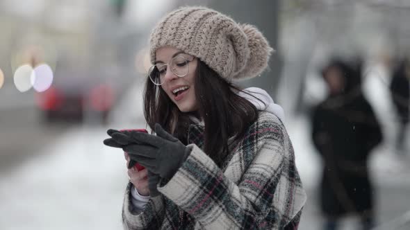 A Beautiful Young Darkhaired Woman in Round Glasses and a Plaid Coat Walks Around the City on a