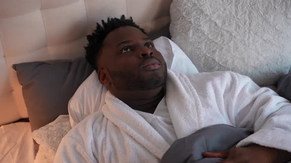 Portrait of Sad Disturbed Young Man Closing Ears with Hands Covering with Blanket Lying in Bed at