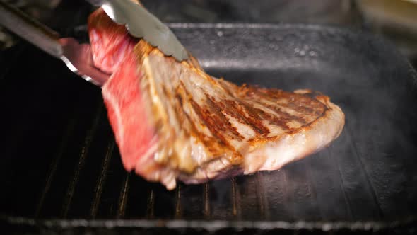 A Piece of Raw Meat Turn By Metal Tongs on a Hot Grill Pan. Cooking T-Bon Steak Closeup. Delicious