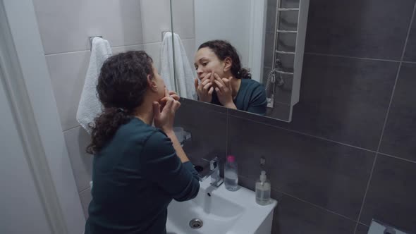 Woman Removing Pimple From Face Looking at Mirror in Bathroom Home