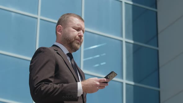 An adult man with stubble in a solid suit and tie is typing on his phone and smiling.