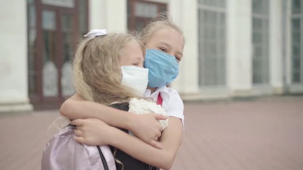 Close-up of Two Schoolgirls in Covid-19 Face Masks Hugging Outdoors. Portrait of Happy Caucasian