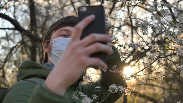 Man in Medical Mask Takes Selfie on Smartphone To Blossoming Apple Tree at Sunset, Coronavirus