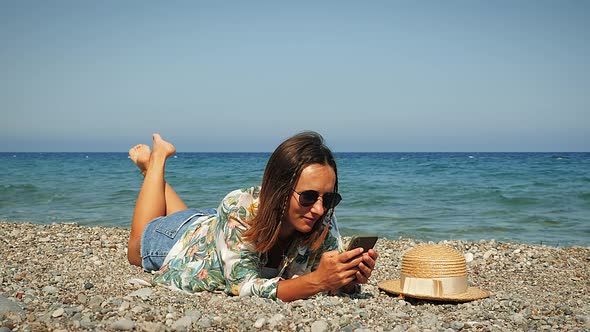 Cheerful charming brunette girl lying on beach and using apps on smartphone at windy summer day. 