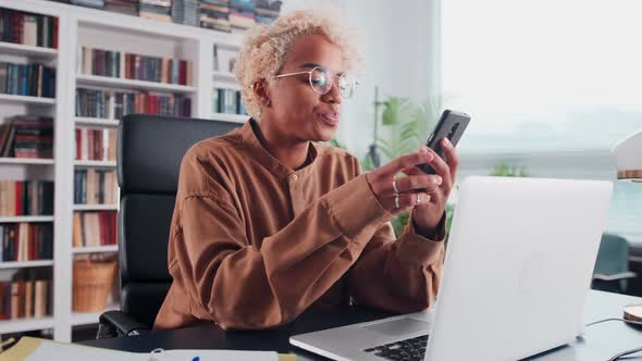 Young African American Business Woman Holding Mobile Phone Sits at Office Desk