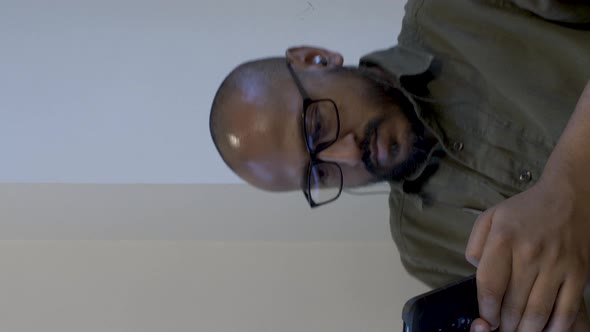 Bald Male Wearing Glasses Holding Smartphone Swivelling In Chair. Vertical Video, Static Shot