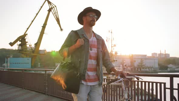 A Young Stylish Hipster with Hat and Glasses Walking with His Bicycle on Sea Port Background During