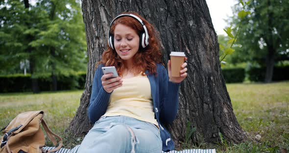 Smiling Girl Listening To Music in Headphones Using Smartphone Relaxing in Park