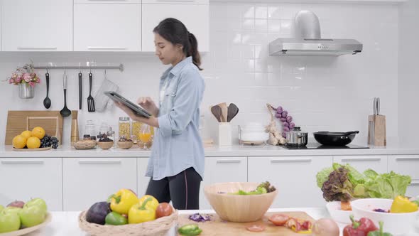 Happy Asian woman using tablet for looking recipe while making food in the kitchen