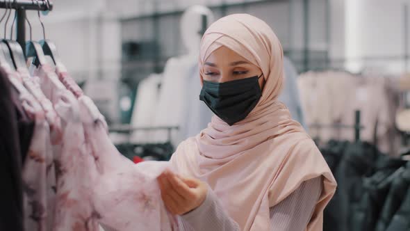 Young Arab Woman Customer Buyer in Hijab Wearing Protective Medical Mask in Clothing Store Carefully