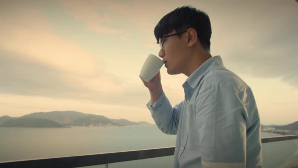 Handsome Dreamy Asian Middle Aged Man Stands in the Balcony with Great Views and Drinks Hot Coffee