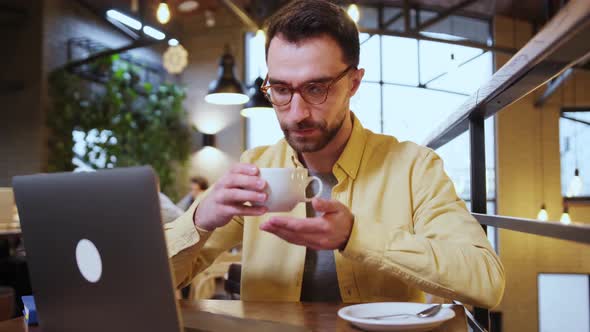 Handsome Man is Enjoying the Aroma of Coffee and Smiling While Working with a Laptop in Cafe