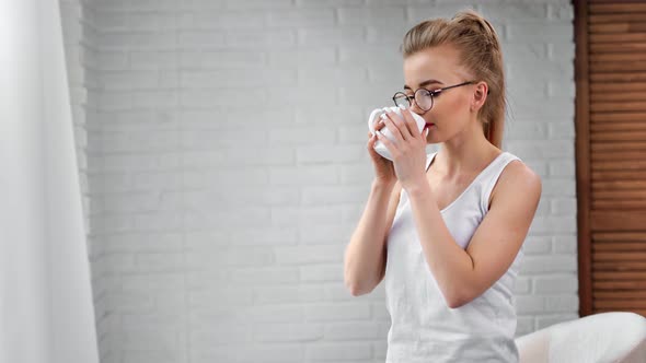 Beautiful Young Domestic Girl in Trendy Glasses Drinking Hot Beverage