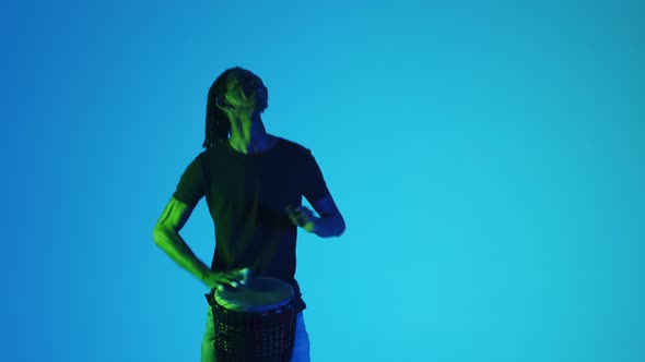 Black Male Musician with Dreadlocks Vigorously and Cheerfully Plays Bongo in Studio Illuminated By