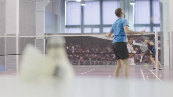 Wide Shot of Two Men Playing Badminton on Indoor Court. Rack Focus Changes To Shuttlecock Lying at
