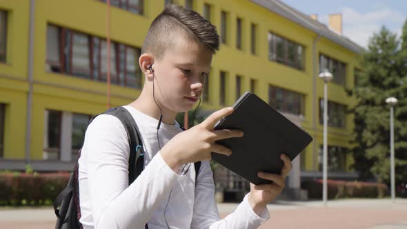 A Caucasian Teenage Boy Plays a Game on a Tablet with Earphones on  a School in the Background