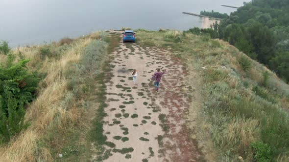 Drone Follows Happy Excited Father and Little Daughter Running to Car on Cliff Over River Bank