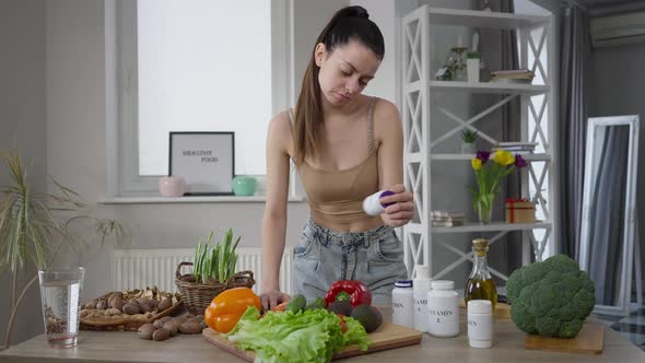 Portrait of Slim Thoughtful Vegan Woman Standing at Table Choosing Vitamins