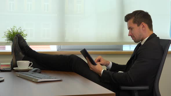 An office worker in a suit sits in front of a computer with legs on the desk and works on a tablet