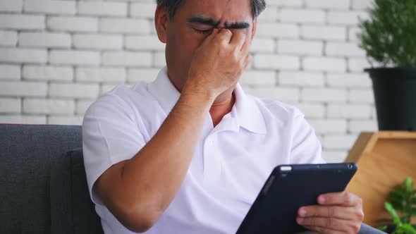 Senior man is suffering from eye strain due to his long-time looking at the digital tablet.