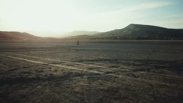 Drone View of Man on Motorbike Rides on a Dry Lake with Dust and Beautiful Hills on Background in