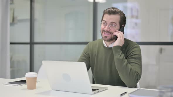 Young Businessman Talking on Smartphone While Using Laptop in Office