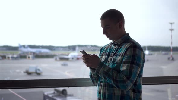 Young Man with a Phone in His Hands on the Background of a Window at the Airport. Airplanes in the