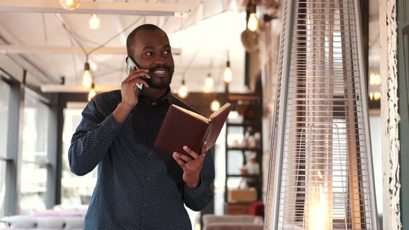 Young African American Man with Book in Cafe and Talking on Phone