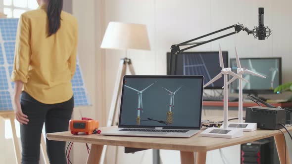 Asian Woman Walks Into The Office That Has Solar Cell Next To The Laptop Showing Wind Turbine