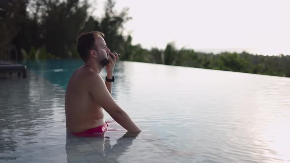 Adult Man Talking on Phone Enjoying Sunset View From Infinity Pool in Rooftop Tropical Hotel