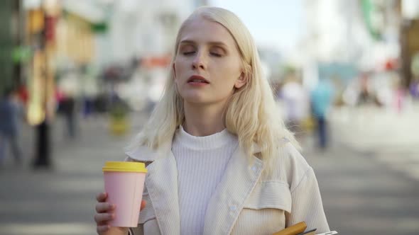 Pensive Young Woman Walks Down the Street with Coffee and Textbooks