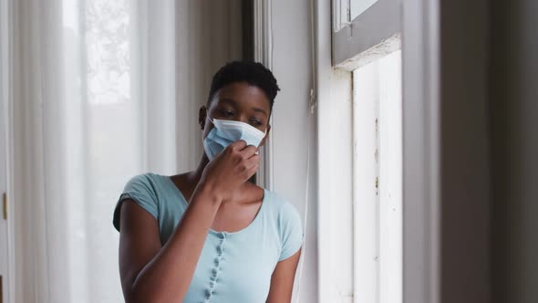 African american woman removing face mask and looking out of window at home