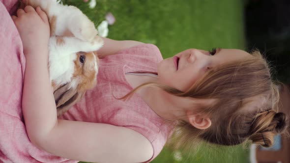 Child Girl in a Pink Dress Hugs an Easter Brown Rabbit Sitting in a Meadow with Green Grass in