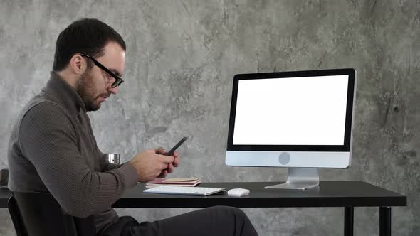 Businessman Looking at His Smartphone Messaging and Sitting Near Computer Screen. White Display