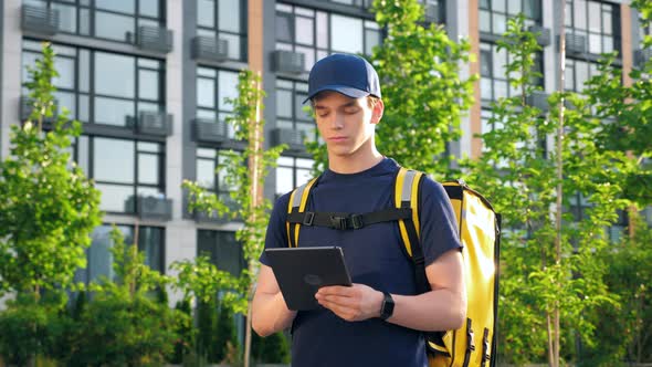 Portrait Young Man Courier Food Delivery with Thermal Backpack Uses Tablet