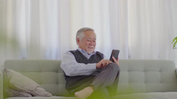 wellness elderly asian man with white hairs sitting on sofa enjoy and using mobile phone