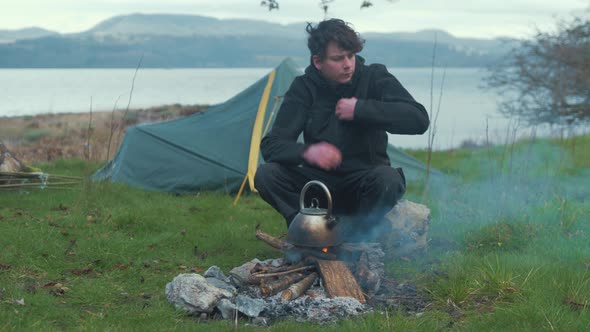 Young man sitting by fire camping on island puts up hood in rain