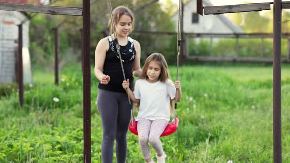 Cheerful Children in Light Clothes Ride a Street Swing in a Blooming Green Home Garden and Laugh in