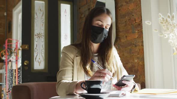 Caucasian female customer wearing face mask, sitting at table, using smartphone