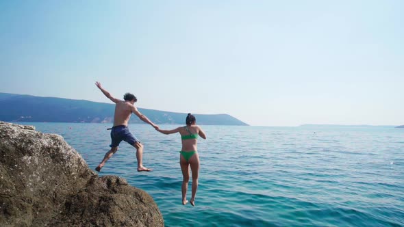 Young couple in swimwear, jumping off from huge rock, into vast blue sea. Slow motion.