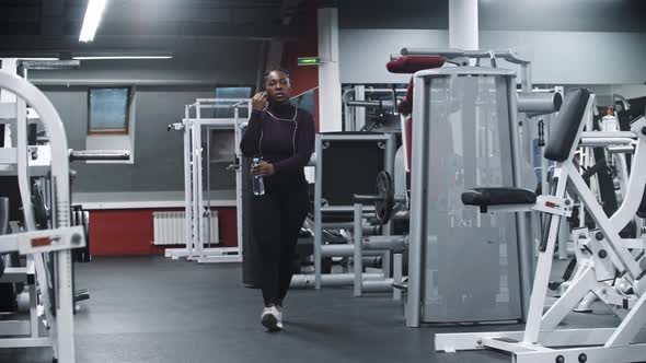 An Africanamerican Woman Puts Headphones in Her Ears in Modern Gym and Drinking Water