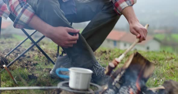 Man Preparing Food, Sitting on Camping Chair in Front of Bon Fire. Barbecue Time in an Autumn Foggy