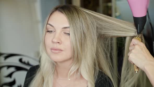 the Hairdresser's Hands are Passing a Strand of Long Blond Hair to Brushing and Hair Dryer