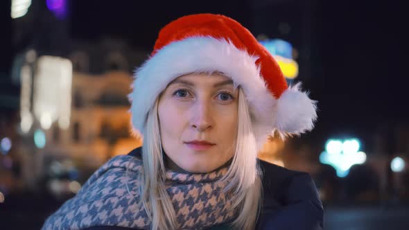 A young woman in a Santa hat looks into the camera . night city.