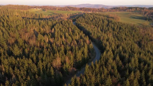 Aerial tracking shot of a white car driving along a curved forest road at sunset. Wide angle drone f