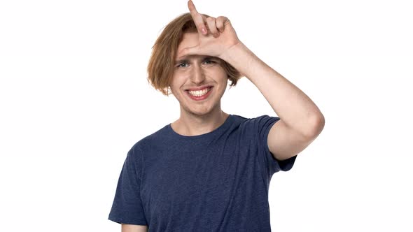 Portrait of Sarcastic Young Guy 20s Grinning and Showing Loser Sign on His Forehead Isolated Over