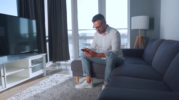 Man with Glasses Sitting on the Couch in a Cozy Room and Using Smartphone for Surfing Internet