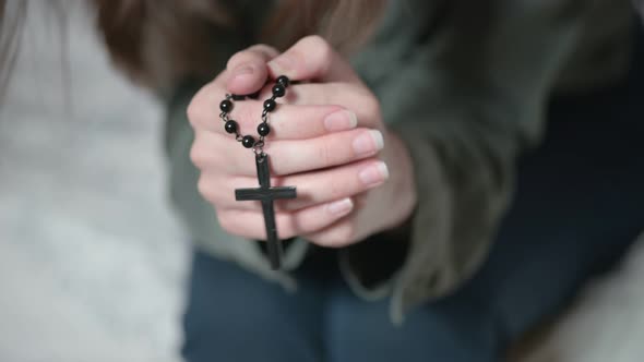 Depressed lady brunette sits on grey sofa and prays holding black rosary in hands and kissing cross