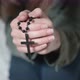 Depressed lady brunette sits on grey sofa and prays holding black rosary in hands and kissing cross - VideoHive Item for Sale